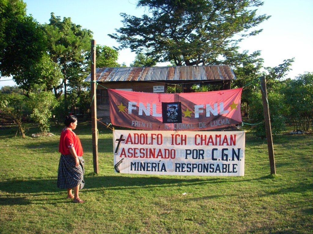 Angelica Choc stands next to a sign that says “Adolfo Ich murdered by CGN. Mining is responsible.” Photograph by Daniel Sosa